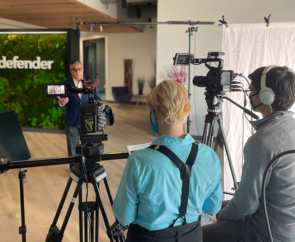 On set of corporate video production of man being interviewed by woman in blue shirt while videographer with headphones manages camera. In corporate office of san antonio company BitDefender.